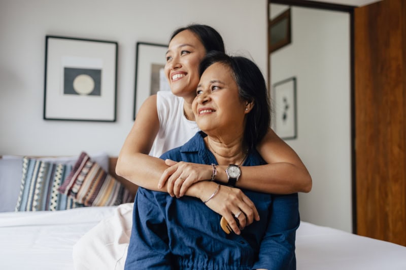a smiling Asian female embracing her mother while they are looking away.